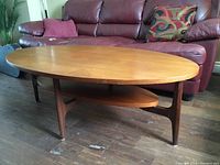 Angled front view of teak oval coffee table with lower shelf and tapered legs
