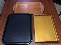 Top view of the three trays comprising one teak tray with handles, one black plastic cafeteria tray, and one gold glitter tray.