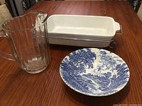 White ironstone casserole dish in chrome rack, clear ribbed glass pitcher, blue and white scenic serving bowl on table.