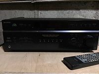 Front view of Sony STR-DE597 digital audio/video control center receiver showing control buttons, display, headphone jack, and remote control on shelf