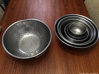 Large stainless steel colander next to stack of eight stainless steel bowls of varying sizes on wooden surface.