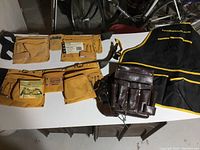 Top-down photo showing all four items on a white table: two suede hip tool belts, one vintage leather pouch belt, and one black Busy Bee apron