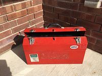 Closed red metal tool box sitting on a concrete floor near brick walls showing the exterior and carrying handle