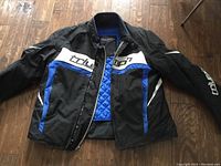 Front view of black Triumph motorcycle jacket with blue and white accents, partially unzipped showing the blue liner inside.