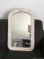 Front view of vintage rectangular mirror with arched top in distressed white wood frame sitting against dark couch.