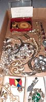 Top-down photo of a box filled with assorted vintage costume jewelry including necklaces, brooches, pins, and a small wood-tone Yellowstone souvenir item. Jewelry shows metal, faux pearl, and rhinestone components.