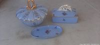 Three-piece blue glass antique vanity set on table, showing lidded bowl, rectangular box with lid, and oval tray, all decorated with floral motifs and gold trim