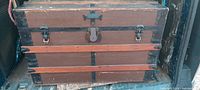 Exterior front view of large rectangular wooden steamer trunk with metal reinforcements and wooden slats. Central lock and two latches visible; leather handles missing.