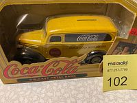 Front view of the yellow Coca-Cola die-cast metal bank in original packaging with visible coin slot on the roof and Coca-Cola branding on the side.