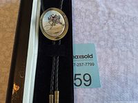 Full view of Disney bolo tie in original black case with white interior lining, showing black braided cords and metal tips alongside the oval slide with Disney illustration.