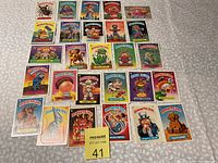Photo of 27 Garbage Pail Kids trading cards laid out on a flat surface showing varied characters and card border colors.