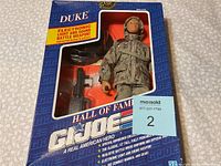 Photo of unopened Hall Of Fame GI Joe Duke action figure in original packaging with visible figure and electronic weapon.