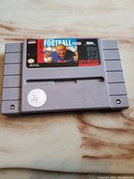 Front view of the grey SNES cartridge showing the John Madden Football label featuring John Madden in the center with NES and Super Nintendo logos.