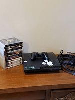 Photo of the PS3 console with controller on top and a stack of 14 PS3 game cases nearby, along with bundled cables.