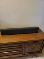 Front view of the Reel RS Cinema center speaker placed on wooden furniture, showing the elongated rectangular black speaker grille.