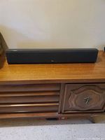 Front view of black Reel RS Cinema 3 channel speaker placed on wooden surface against wall.