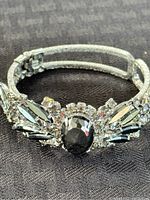 Front view showing silver-tone bracelet with central large black oval stone and surrounding clear and black rhinestones in floral pattern.