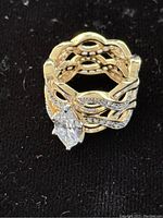 Front view of gold colored ring showing the large clear marquise-cut stone and intricately designed wide band with small stones embedded.