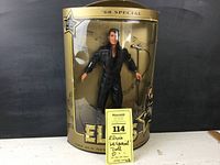 Front view of the Elvis '68 Special doll dressed in black leather inside original packaging missing top cover, with auction tag visible.