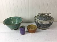 Four modern art pottery pieces shown together: large green bowl, two smaller speckled vessels in purple and orange, and a large lidded steamer with detailed floral and geometric design and handle.