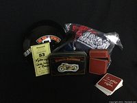 Photo showing lot contents: black novelty half helmet with colorful Daytona Harley Davidson stickers, sealed bag of assorted 100% cotton bandanas, Harley Davidson metal tin with 1997 playing cards, and brown leather carrying case with playing cards.
