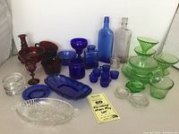 Wide view showing mixed vintage glass pieces in cobalt blue, deep ruby red, clear, green glass including bowls, plates, bottles, and cups.