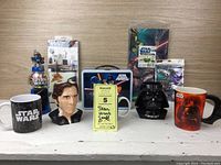 Star Wars mugs including Darth Vader, Han Solo bust, and other character-themed mugs along with Star Wars wall decals, stickers, and M&M dispenser shown on a white surface