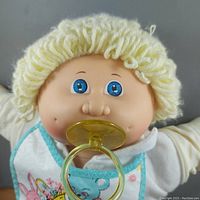 Close-up front face view of the doll showing blue eyes, blonde yarn hair, and yellow pacifier in mouth, wearing embroidered bib