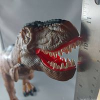 Close-up photo of the dinosaur toy's head showing textured skin, open mouth with red gums and white teeth, positioned next to a ruler indicating length about 11 inches.
