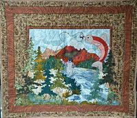 Photo of a quilted fabric tapestry approximately 30 x 24 inches depicting a fly fishing scene with a fisherman, fish, trees, mountains and water, bordered with wood grain and leaf patterned fabrics.