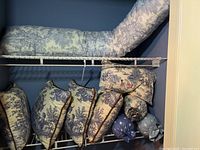 Nine decorative pillows arranged on wire shelving in a closet area, showing various shapes and sizes in blue and white toile fabric with pastoral and countryside scenes.
