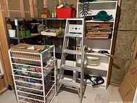 Overview of metal wire rack with multiple shelves showing assorted contents such as boxes, a step stool, and a 5-foot aluminum ladder behind it in basement setting.