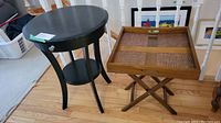 Round black wood table with drawer and bottom shelf and folding wooden table with removable tray top positioned next to railing on wood floor.