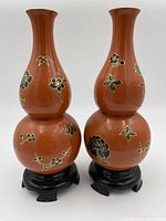 Pair of Asian lacquer double gourd vases with black and gold floral designs on reddish-brown lacquer finish, displayed on black lacquered wooden stands.