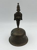Front view of vintage bronze bell with standing figure handle and engraved floral designs on the surface.