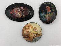 Photo showing three lacquer enamel brooches and pins with hand-painted floral, rural house, and fishing man designs on dark lacquer backgrounds.