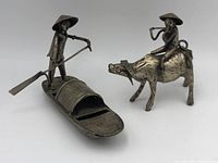 Photo showing two metal statues: one man rowing a woven-pattern boat, the other man sitting on water buffalo playing flute