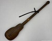 Wooden paddle-shaped item with a long handle, black cord looped through hole at handle end, tied string near middle, showing signs of wear.