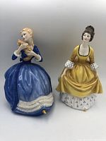 Two Royal Doulton porcelain doll figurines placed side by side on a white background. One doll in a blue dress holding a cat, the other in a yellow dress.