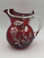 Front angle showing the sterling silver floral overlay on deep red glass jug with clear handle and silver rim