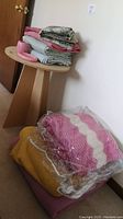 Stack of vintage blankets with pink and white crochet afghan on bottom, and yellow blanket below along with pressboard side table holding folded fabric and accessories.
