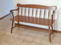 Front view of a colonial style solid oak bench showing the seat, backrest with vertical spindles, armrests, and turned legs.