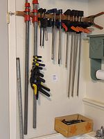 Wide shot of multiple clamps hanging on wall-mounted wooden rack, showing long bar clamps, smaller quick-release clamps, and spring clamps.