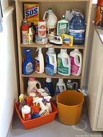 Full view of shelving unit with various cleaning supplies including SOS pads, Sunlight soap, Quick Shine, multiple spray bottles, and orange bucket and tub with contents on floor.