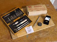 Full view of all items laid out on wooden cutting board including cutting kit case, electric engraver box, measuring tape, tie pin in box, and cutting board.