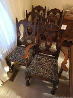 Photo showing one armchair and five side chairs with carved wood backs and floral upholstered seats, arranged near a window and sideboard.