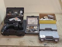 Lot overview showing cordless drill on black case, power drill, hole saw kit, sanding drum kits in plastic and cardboard boxes