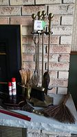 Full view of brass fireplace tool set in stand with brush, scoop, poker, tongs and additional items including matches, lighters, and sweep items set on fireplace hearth against brick wall.