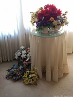 Full view of the round composite wood table with glass top, covered by cream ruffled tablecloth, with multiple artificial floral arrangements on and around it.