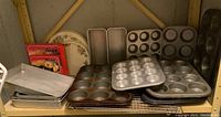 Wide view of multiple muffin tins, cake loaf pans, cooling racks, and decorative stove burner covers arranged on a shelf.